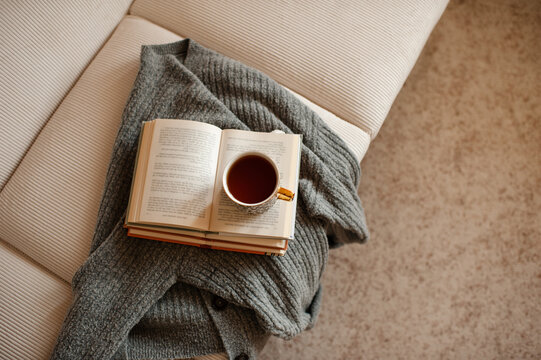 Cup of black tea staying on open paper books with knitted textile on white velvet couch  in living room at home. Cozy and hygge atmosphere. Coziness.
