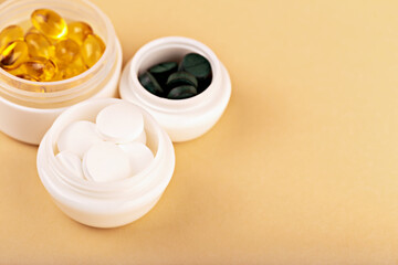 Close-up of white cosmetic containers filled with yellow capsules, green spirulina and white tablets on beige background. Concept of vitamins and natural health care. Various supplements in white jars