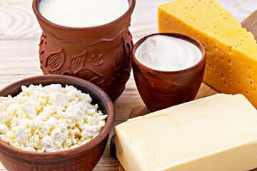 Rustic dairy assortment with milk in clay jug, cottage cheese, sour cream, butter and slice of yellow cheese arranged on wooden table. Farm-style selection highlighting fresh and natural dairy product