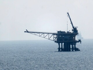 Offshore oil platform dominates the image, situated in a calm ocean under a hazy sky. A distant ship is visible. The background is a solid, hazy blue. The platform appears still, 