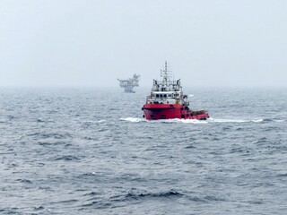 A red tugboat navigates the vast ocean, a distant oil rig barely visible under a hazy sky. The waves gently rock the vessel, creating a tranquil yet powerful scene of maritime industry.