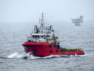 A powerful red tugboat cuts through the ocean waves, heading towards a distant offshore platform on a misty day. The vessels movement creates dynamic splashes and froth.