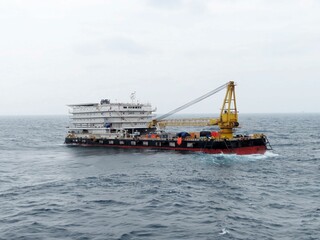 A large barge with a yellow crane sails across a dark blue ocean under a pale gray sky. The vessel appears sturdy and functional, navigating choppy waters. Its a scene of maritime industry at work.