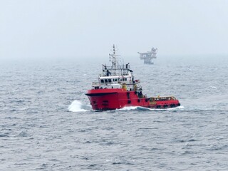 A vibrant red tugboat navigates the choppy ocean waters, a distant offshore oil rig barely visible under a hazy sky. The vessel cuts through waves, showcasing its power and resilience at sea.