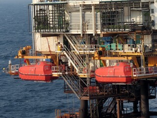 Two red life rafts hang from an offshore oil platform above a dark blue, choppy ocean. The platform shows signs of rust and wear. The overall atmosphere is one of industrial activity.