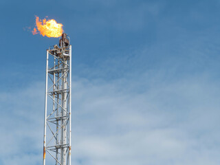 A metal tower with a bright flame burning at the top is set against a clear blue sky with light clouds. The tower appears tall and slender. The background is solid, not transparent. 