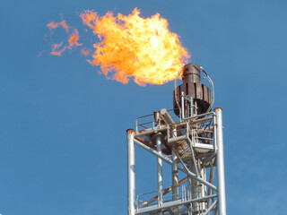 A large orange flame erupts from a metallic flare stack against a clear blue sky. The stack is tall and appears weathered. The background is a solid, unblemished blue, suggesting a daytime setting. 