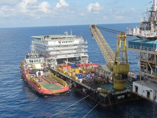 A massive offshore platform receives supplies via a specialized barge and crane, a testament to the intricate logistics of deep-sea operations under a partly cloudy sky.