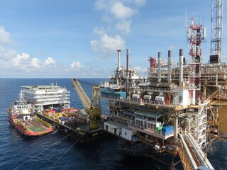 A large offshore oil platform dominates the image, with a support vessel alongside. The ocean is a deep blue, the sky is bright. The platform shows intricate structures, conveyors and equipment. 