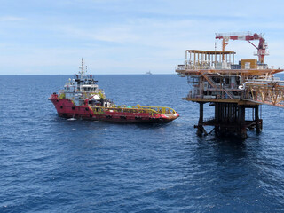 A red tugboat approaches a large offshore oil platform under a clear blue sky. The calm ocean reflects the sunlight, creating a serene yet industrial scene. Distant vessels dot the horizon.