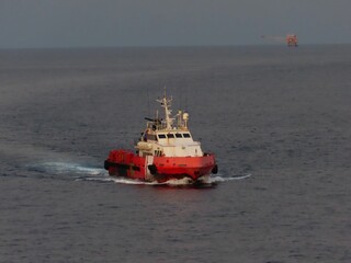 A vibrant red tugboat cuts through the deep blue sea, leaving a frothy wake. In the distance, an oil rig stands silhouetted against the dusky sky, a solitary sentinel in the vast expanse of water.