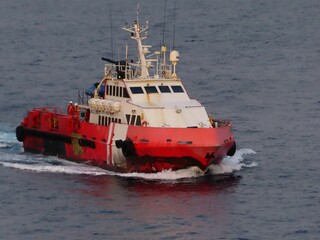 A powerful red supply vessel cuts through the dark blue ocean waters, its wake a frothy white trail. Crew members are visible on deck, navigating the open sea under a calm, dusky sky.