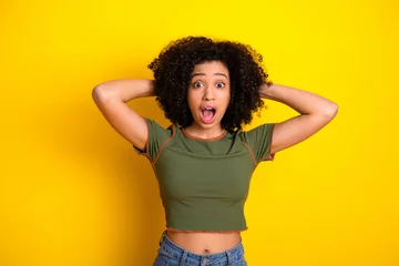 Gordijnen Surprised young woman with curly hair making a shocked expression against a bright yellow background © deagreez