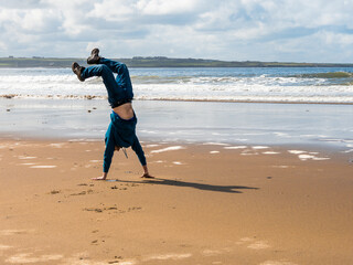 Energetic man doing hand stand on a sandy beach, blue picturesque sky in the background. Concept of freedom and power, as the man shows his ability in a public place. Sport and fitness.