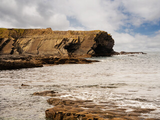 A rocky cliff coastline by the ocean. The water is calm and the sky is overcast. The scene is...