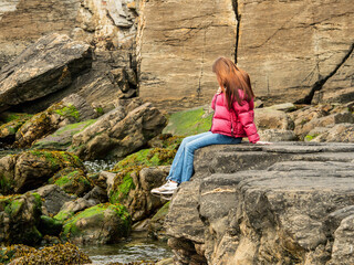 A woman sitting on rough stone coast. She is wearing a red jacket and blue jeans. The scene is...