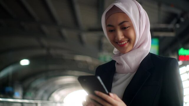 Happy asian muslim business woman wearing hijab holding and using smartphone waiting railway or skytrain at train station. Islamic woman people lifestyle travel. Muslim people concept.
