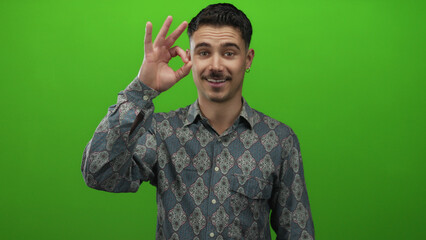 Young hispanic man in patterned shirt isolated against vivid green background making okay gesture...
