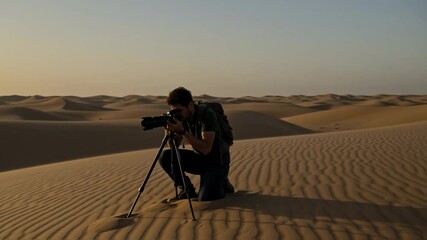 Photographer with a backpack and camera on a tripod captures the golden sunset amidst vast desert sand dunes, embodying adventure landscape photography and creative exploration in remote nature