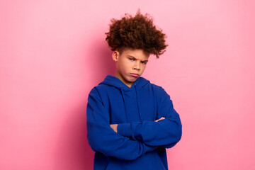 Cute young boy in blue hoodie with arms crossed standing against a pink background, expressing...