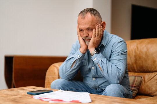 Man sitting on a couch in a living room looks stressed while examining papers on a wooden table during the afternoon