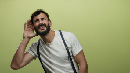 Hispanic man with beard listening intently against a yellow backdrop wearing a white shirt and...