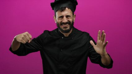 Hispanic bearded man chef gesturing aggressively in black uniform against a vibrant pink background portraying frustration and emotion.