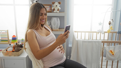 Pregnant woman with long blonde hair sits on bed in cozy bedroom, smiling at her phone surrounded by baby toys and a cradle, creating a warm, family-oriented atmosphere.