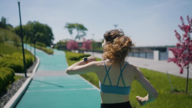 Back view of a young sportswoman with curly hair, wearing a blue sports bra and black leggings, running on a green track in a city park, checking her smartwatch during her training on a sunny day