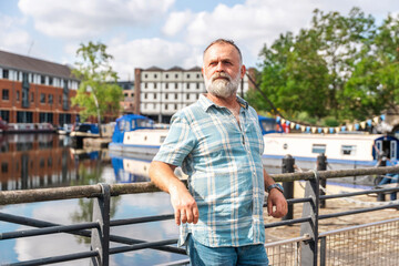 Bearded man poses confidently by the water in a lively urban marina during a sunny day with greenery in the background