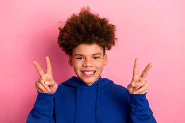 Cheerful young boy in blue hoodie making peace gestures against pink background