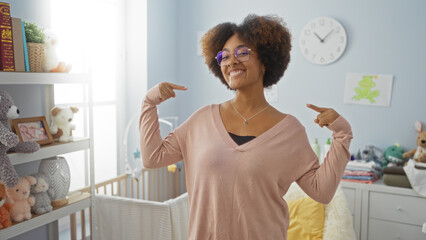 Woman smiling with curly hair points to herself in a cozy bedroom with a baby cradle surrounded by soft toys and pastel decor, radiating warmth and style.