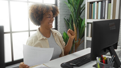 Woman celebrating success in modern office with documents and computer, surrounded by bookshelves and plants, showcasing an inspiring professional environment.