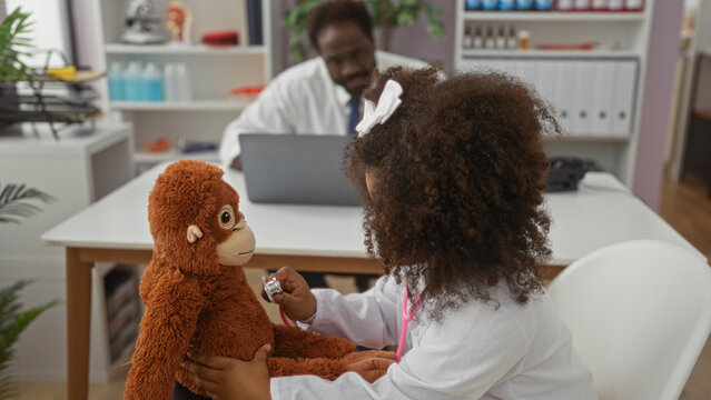 Girl playing with teddy in a clinic room with father working on laptop nearby depicting joyful family interaction in healthcare environment