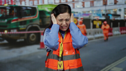 Woman volunteer covers ears in noisy construction site wearing orange safety vest and blue sweater...