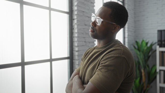 Man standing in modern office room with natural light, wearing glasses and a casual shirt, exuding confidence and focus, plant and shelves in the background.