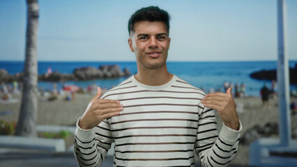 Handsome young man gesturing in a striped shirt at a vibrant seaside beach with palm trees and ocean in the background, conveying a casual summer vibe outdoors.