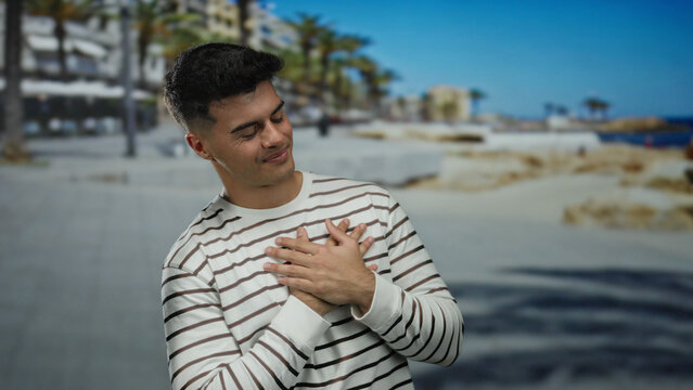 Young man in striped shirt at beach expressing contentment with eyes closed, hands on chest, outdoors beside the ocean, amid coastal landscape and palm trees - Powered by Adobe