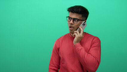 Young man in glasses talking on phone over isolated green background, wearing red sweater,...