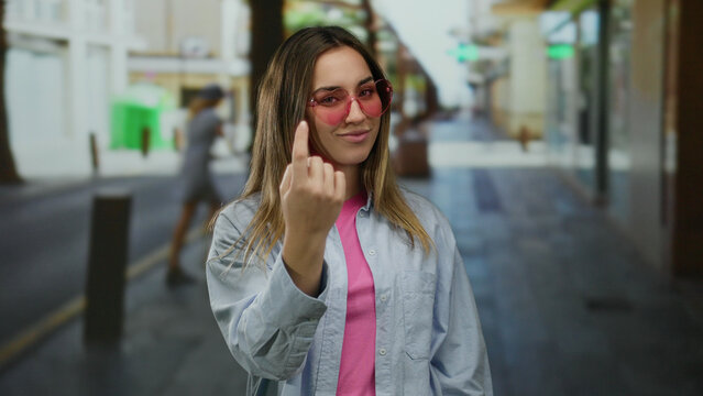 Woman smiling and gesturing come with her finger on a sunny street wearing sunglasses and a casual shirt showcasing a playful inviting demeanor in an urban outdoor setting.