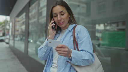 Fototapeta premium Woman on urban street talking on phone, wearing blue shirt, holding card, outdoors, daytime, looking focused, carrying bag, standing near building reflection.