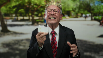 Senior man in business suit with crown laughing in park, showcasing joy with credit card in hand...