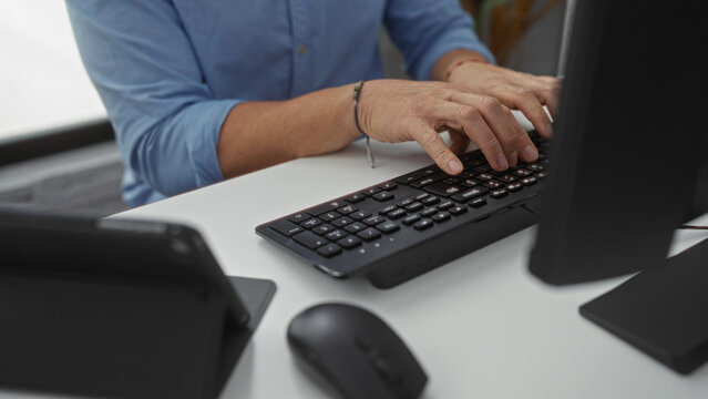 Man typing on keyboard in modern office setting with focus on hands and technology, wearing casual blue shirt, busy in indoor workplace environment.