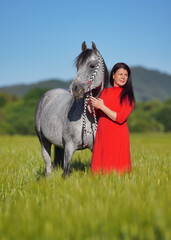 Woman with dark hair and bright red dress walking her Arabian gray horse in green field on sunny day, smiling as animal eats grass