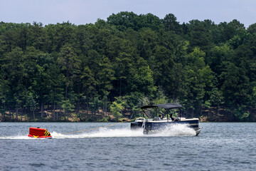 Fototapeta premium Pontoon boat pulling inflatable tube float raft on lake. Family on a pontoon party boat enjoying tubing on a summer day with their children.