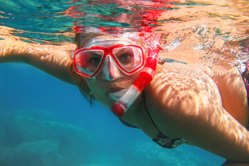 Young woman in red diving mask and snorkel swimming in a sea looking into camera, underwater photo