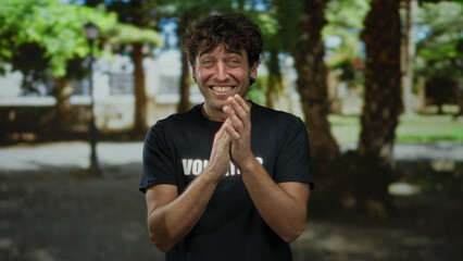 Hispanic man clapping joyfully in a green park wearing a volunteer shirt, exuding charm and...