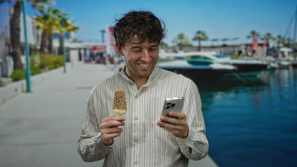 Hispanic man enjoying ice cream while checking smartphone at the seaside marina on a sunny day.