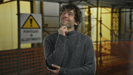 Young man thinking indoors at construction site wearing sweater with thoughtful expression near warning sign background.