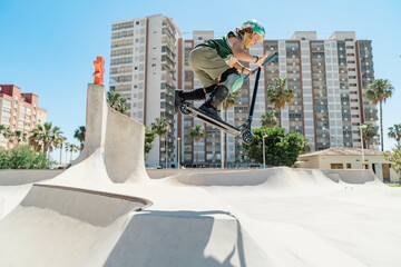 Young scooter rider performing stunt in skatepark with buildings in background